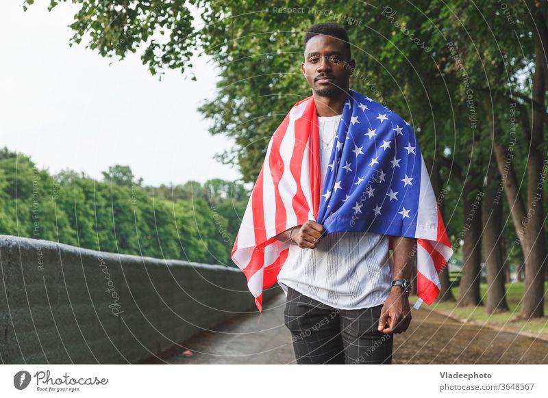 Gutaussehender afroamerikanischer Mann mit der USA-Flagge auf den Schultern, der ernsthaft in die Kamera schaut und im Freien steht. Tag Sommer Amerikaner
