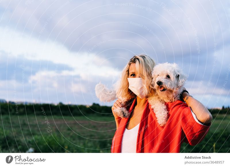 Zufriedene Frau mit Atemschutzmaske und niedlichem kleinen Hund auf den Schultern Bichon frise Atemschutzgerät Landschaft Natur lustig Coronavirus Inhalt lässig