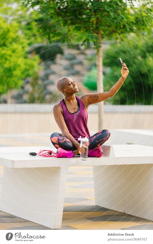 Fröhliche schwarze Sportlerin, die sich nach dem Training am Smartphone selbstständig macht Selfie Pause Sommer Großstadt Sportbekleidung Bank Flasche benutzend