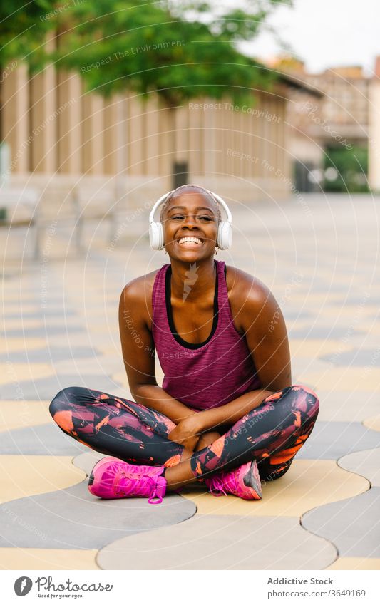 Glückliche schwarze Sportlerin mit Kopfhörer sitzt nach dem Training auf dem Bürgersteig Athlet Headset Sportbekleidung Stauanlage heiter nach vorne lehnen