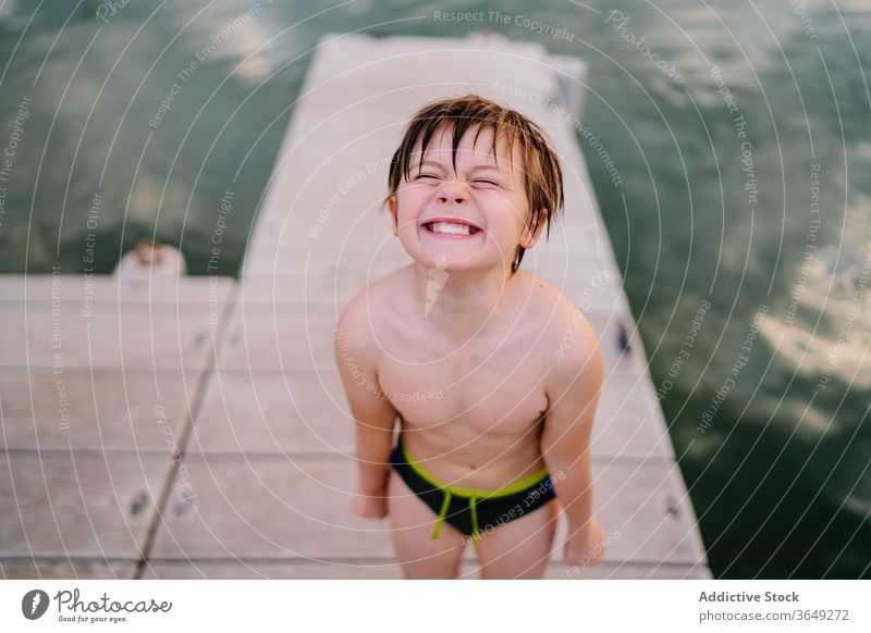 Entzückter Junge auf der Mole am See Gesicht machen Lächeln heiter Pier Badebekleidung Kai lustig Grimasse Glück Freude Kind Kindheit Teich Wasser Sommer ruhen