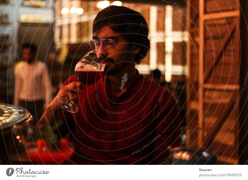 Junger Mann mit rotem Hemd, während er Rotwein aus einem Kelch trinkt, in einer Bar mit bunten Lichtern um ihn herum. Lifestyle trinken im Innenbereich Feier