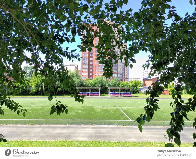 Leeres grünes Fußballfeld in der Nähe einer Schule und des Wohnhauses in Russland. Stadion und Fußballfeld mit leeren Zuschauerplätzen. Fit halten und sich im Freien bewegen. Stadtansicht in einem Rahmen aus Blättern.