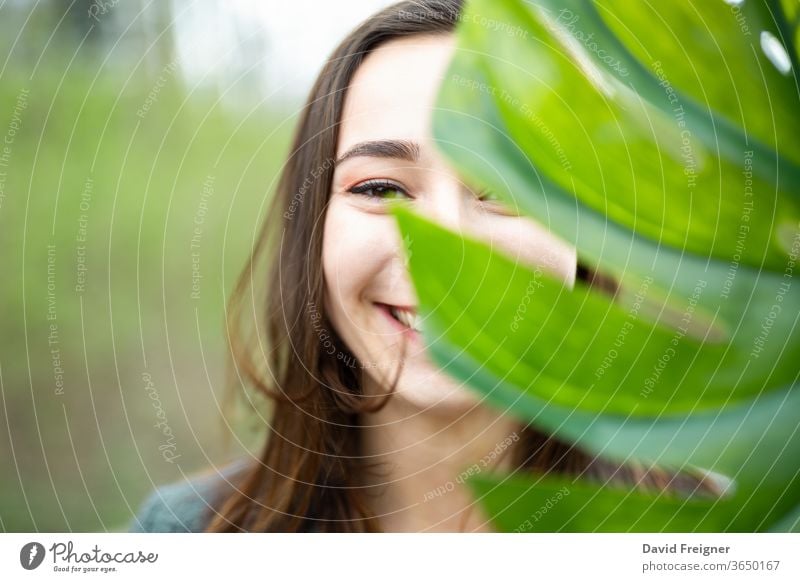 Schöne natürliche junge Frau in Nahaufnahme hinter einem großen Monsterblatt mit grünem Hintergrund in den Wäldern. Lächeln Gesicht Blatt attraktiv Sauberkeit