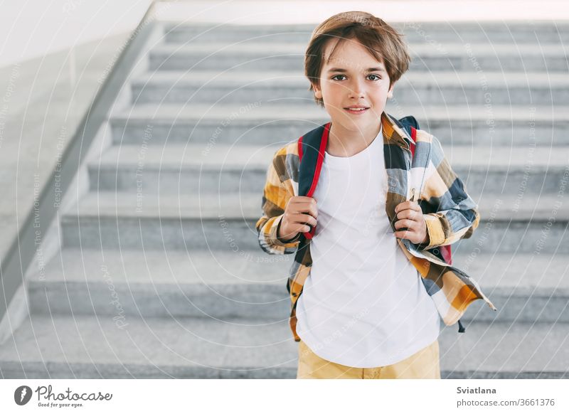 Ein glücklicher Junge mit einem Rucksack steht auf den Stufen vor dem Eingang der Schule und lächelt wunderschön. Beginn des neuen Schuljahres nach den Sommerferien. Zurück zur Schule