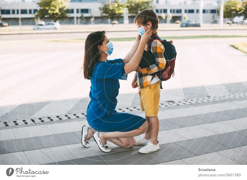 Mama setzt einem Kind eine Maske auf und begleitet es zur Schule oder in den Kindergarten. Ein Junge mit einem maskierten Rucksack während eines Coronavirus. Seitenansicht