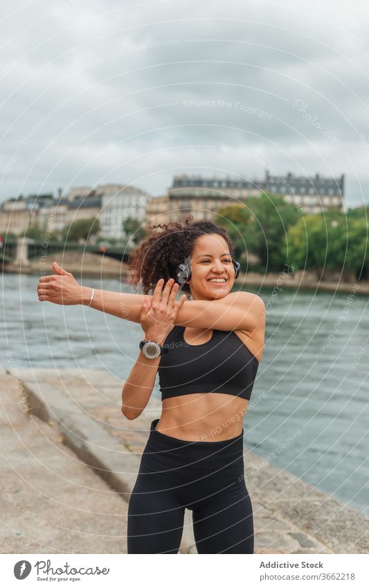 Fröhliche ethnische Sportlerin beim Headset-Training am Stadtfluss Athlet zuhören Musik intelligente Uhr Energie Fluss benutzend Gerät Apparatur Großstadt