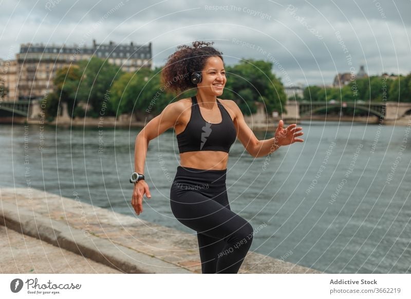 Fröhliche ethnische Sportlerin beim Headset-Training am Stadtfluss Athlet zuhören Musik intelligente Uhr Energie Fluss benutzend Gerät Apparatur Großstadt