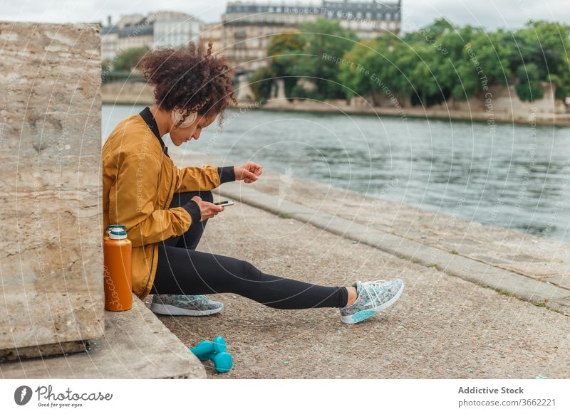 Sportlerin mit Headset nutzt Social Media auf Smartphone nach dem Training Kopfhörer soziale Netzwerke plaudernd Internet Pause Stauanlage Fluss benutzend