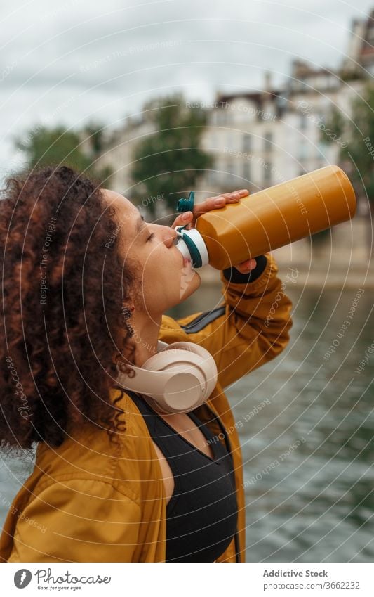 Durstige ethnische Sportlerin trinkt Wasser, nachdem sie in der Nähe eines Flusses trainiert hat trinken Flasche Headset Sportbekleidung Pause Baum benutzend