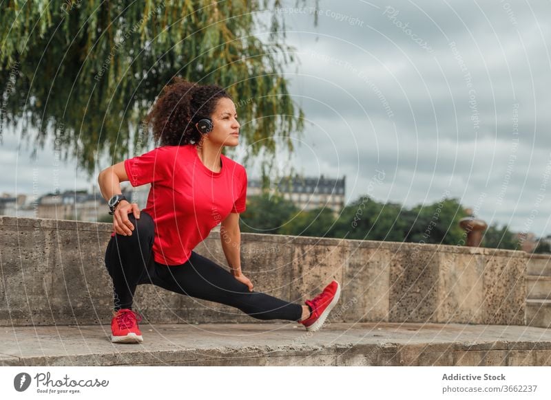 Ethnische Sportlerin mit Kopfhörer, die beim Aufwärmen im Freien ihr Bein streckt Kniebeuge Dehnung Training Übung Drahtlos zuhören Musik Headset beweglich