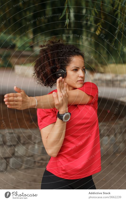 Ethnische Sportlerin mit Headset, die während des Trainings im Park den Arm streckt Athlet Dehnung Arme intelligente Uhr Übung zuhören Musik Drahtlos erreicht