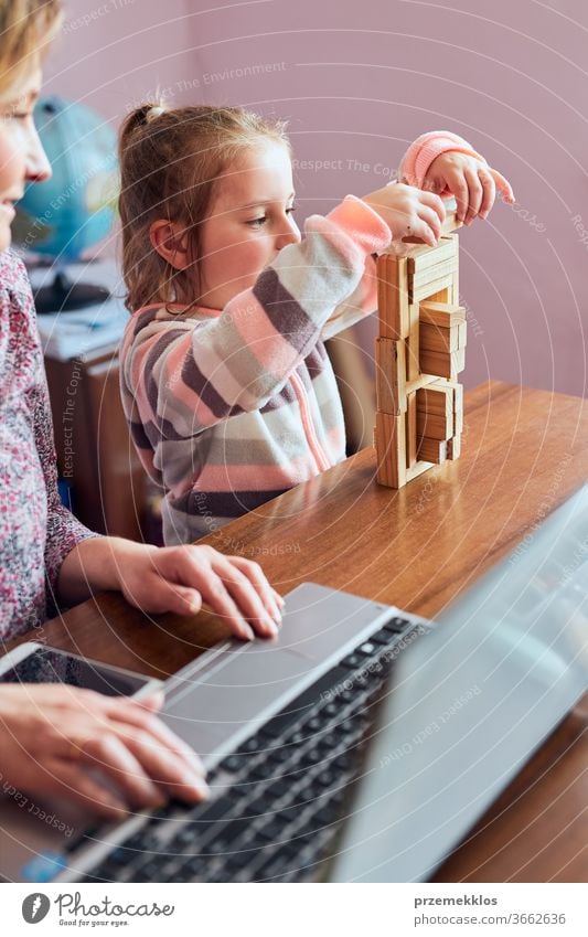 Eine Mutter, die während eines Video-Chat-Call-Stream-Online-Kurses von zu Hause aus auf einem Laptop arbeitet, während ihre Tochter mit einem Ziegelsteinspielzeug spielt. Frau sitzt am Schreibtisch vor dem Computer und schaut auf den Bildschirm