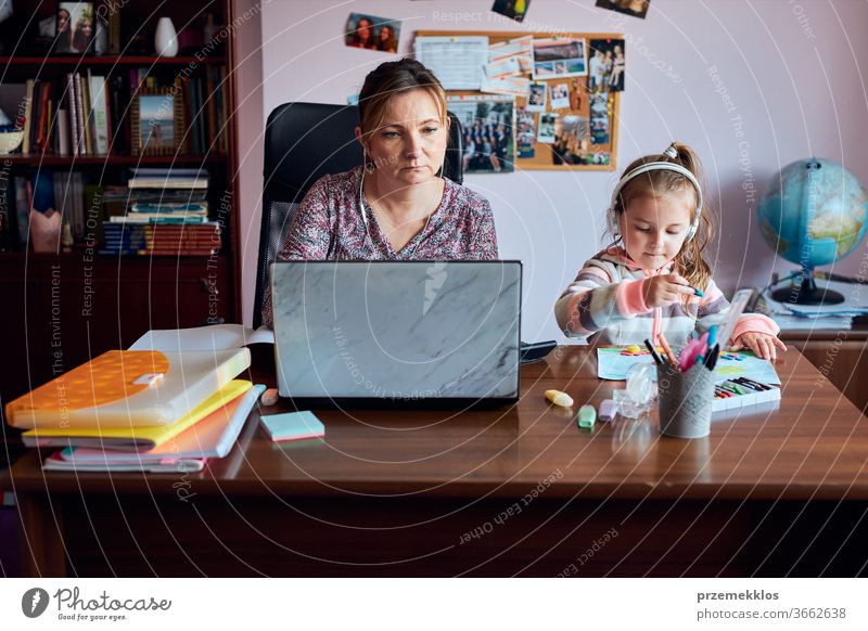 Eine Mutter, die während eines Video-Chat-Call-Stream-Online-Kurses auf einem Laptop von zu Hause aus aus der Ferne arbeitet, während ihre Tochter Zeichnungen anfertigt, herumspielt und sich Geschichten anhört. Frau sitzt am Schreibtisch vor dem Computer und schaut auf den Bildschirm
