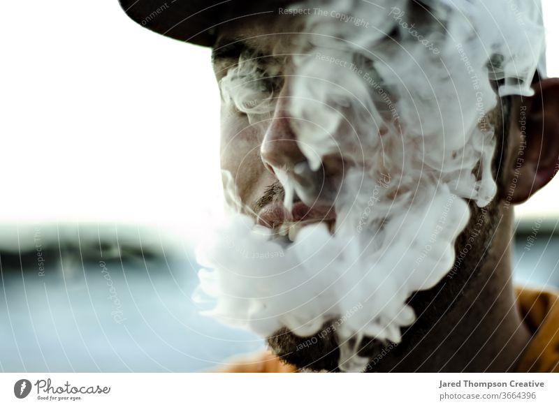 Ein junger, bärtiger Mann bläst dicken Rauch aus seinem Mund aus einem Vape. Raps Rauchen Cloud Jugend Erwachsener Teenager Zigarette Rauchwolke rauchend Sucht