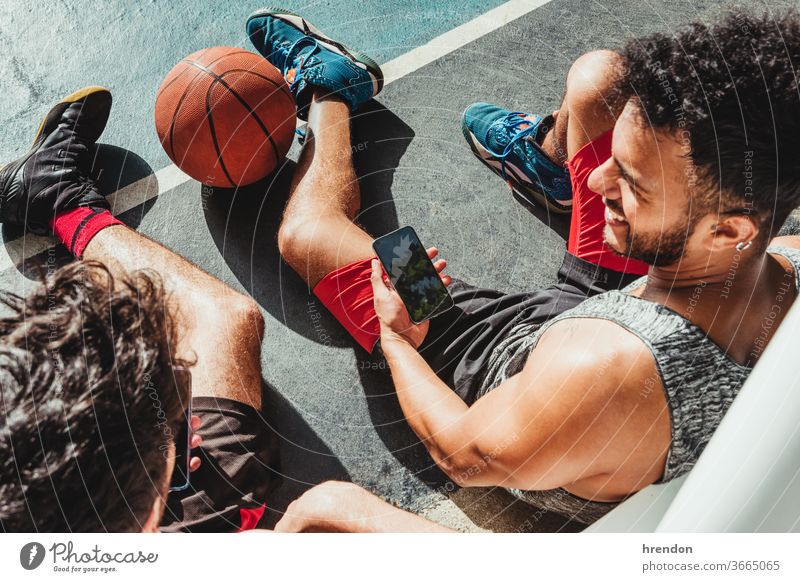 junger Mann ruht sich nach dem Basketballspiel aus und benutzt sein Smartphone mit seinem Freund Sport Ball Spiel sportlich soziale Netzwerke wettbewerbsfähig