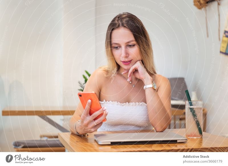 Positive junge Frau benutzt Smartphone, während sie mit Laptop und Saft am Tisch sitzt lesen Nachricht Pause Café Haare berühren besinnlich Gerät Apparatur