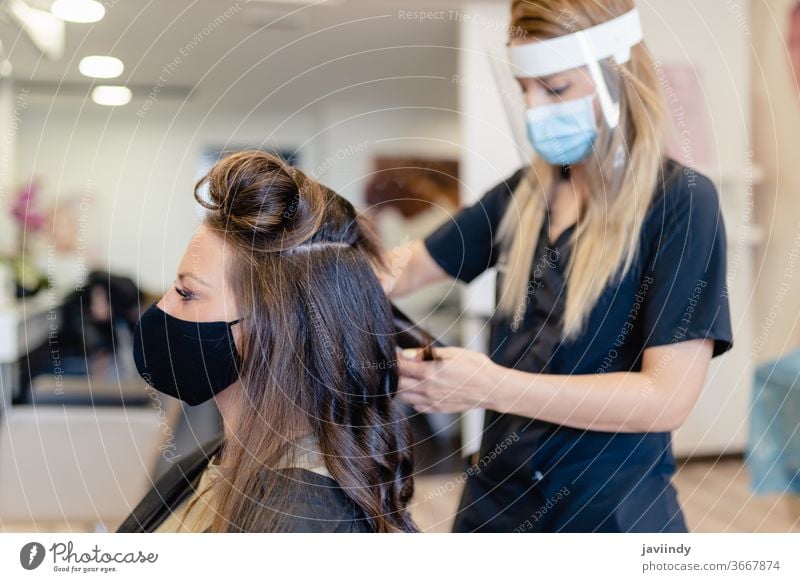 Friseurin, durch eine Maske geschützt, kämmt in einem Salon die Haare ihrer Kundin mit einem Bügeleisen. Frau bügeln Wellen Behaarung Mädchen Frisur Pflege