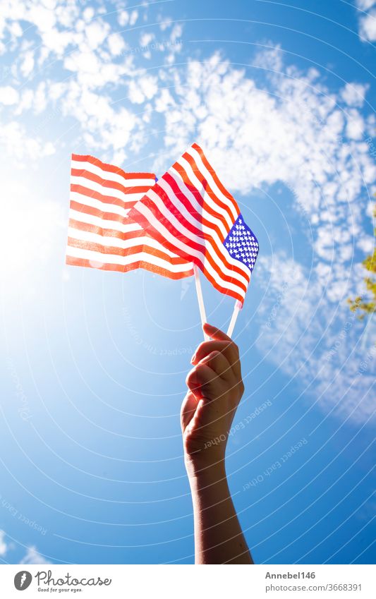 Hand hält zwei amerikanische Flaggen am blauen Himmel mit Sonnenhintergrund, winkende Fahne für die Vereinigten Staaten von Amerika in Nahaufnahme national