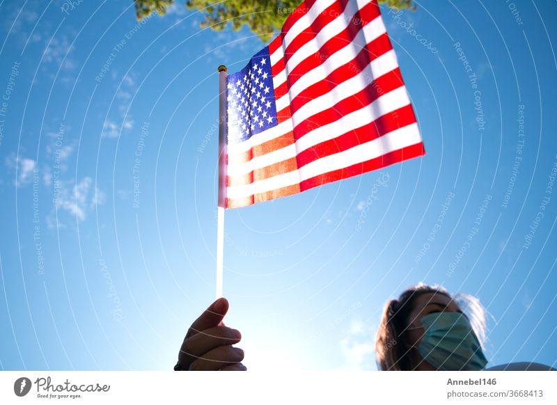 Junge Frau hält amerikanische Flagge am blauen Himmel mit Sonnenlicht und Sicherheitsmaske für Covid-19, der für Usa winkt USA Patriotismus Patriotin im Freien