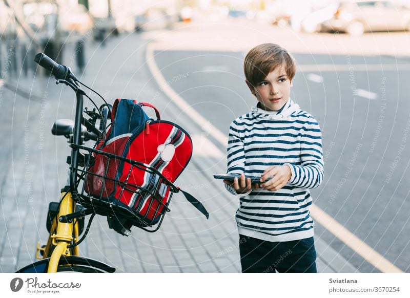 Ein süßer Junge in einem gestreiften Pullover hält eine Tafel in der Hand und steht neben einem Fahrrad, an dem ein Rucksack hängt. Der Junge wird nach der Schule auf einem Fahrrad nach Hause fahren. Sicherer Heimweg