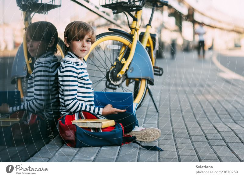 Ein niedlicher Schuljunge sitzt in der Schule unter freiem Himmel und hält ein Buch in den Händen. Daneben ein Schulrucksack und ein Fahrrad, Platz für Text