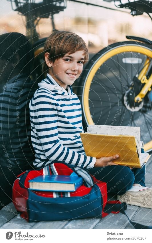 Ein fröhlicher Junge sitzt auf dem Bürgersteig in der Nähe der Schule und liest ein Buch. Daneben steht ein Fahrrad und ein Rucksack. Gesicht müde Deckung