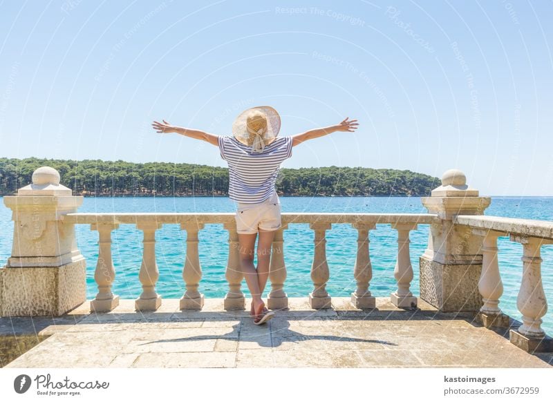 Rückansicht einer glücklichen Frau im Urlaub, die einen Strohhut trägt, auf einem luxuriösen, eleganten, alten Steinbalkon einer Küstenvilla steht, sich entspannt, die Arme zur Sonne erhoben und auf das blaue Adriatische Meer schaut.