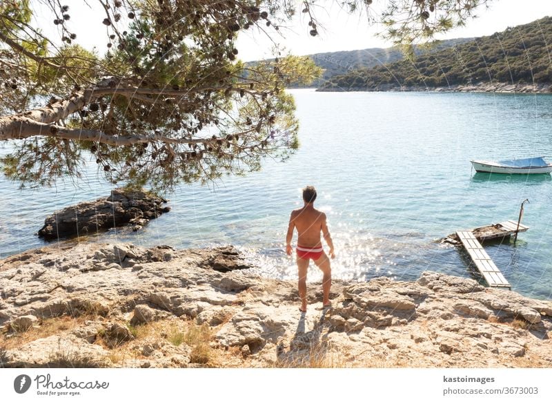 Rückansicht eines Mannes in roter Badehose, der sich an einer wilden Bucht der Adria an einem Strand im Schatten eines Kiefernwaldes sonnen und entspannen will. Konzept einer entspannten gesunden Lebensweise.