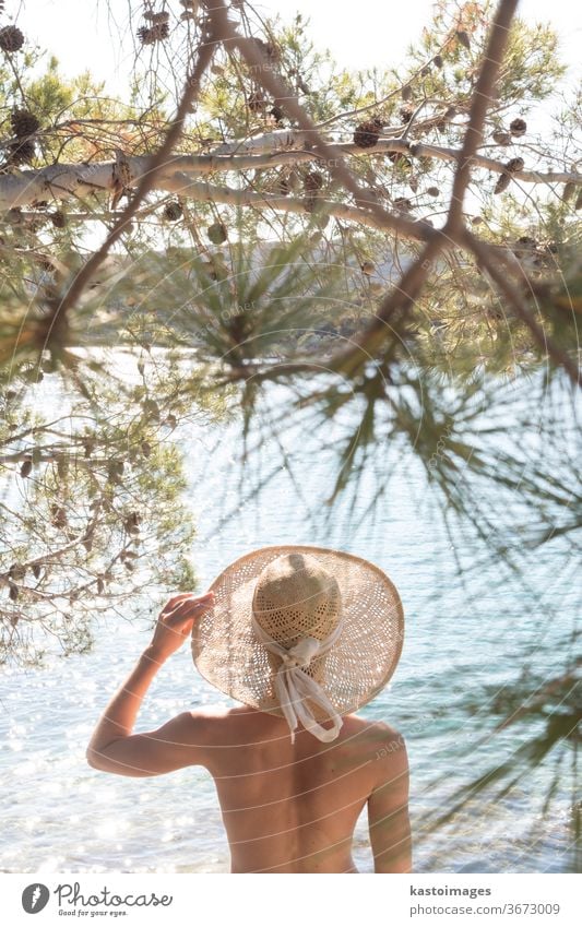 Rückansicht einer oben ohne schönen Frau mit nichts als einem Strohhut, die sich an der wilden Küste der Adria an einem Strand im Schatten einer Pinie entspannt. Entspanntes Konzept eines gesunden Lebensstils.