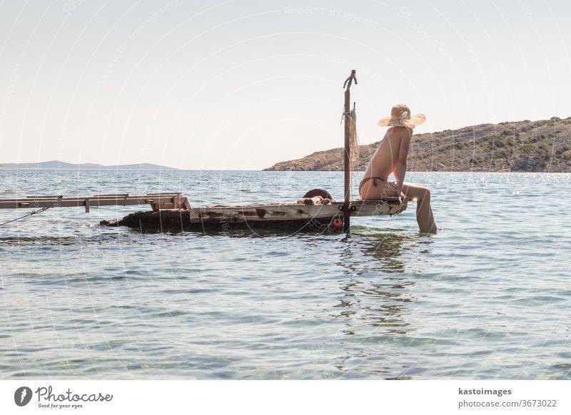 Blick auf eine nicht wiederzuerkennende Frau mit großem Sommersonnenhut, die sich oben ohne bräunt und auf einem alten Holzsteg in einer abgelegenen ruhigen Bucht an der kroatischen Adria entspannt.