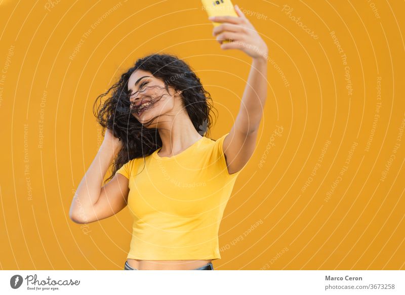 junge gemischtrassige Frau in gelbem T-Shirt, die mit ihrem Telefon mit orangefarbener Wand im Hintergrund einen lustigen Selfie macht unter Smartphone