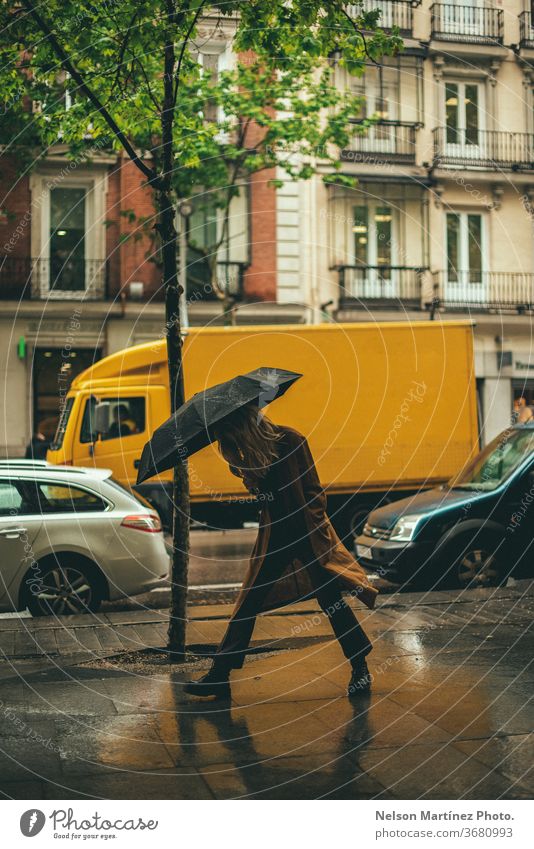 Stilvolle blonde Frau, die im Regen spazieren geht. Sie hält einen schwarzen Regenschirm in einem nicht gesperrten Bereich. regenreich Wetter nass Mensch
