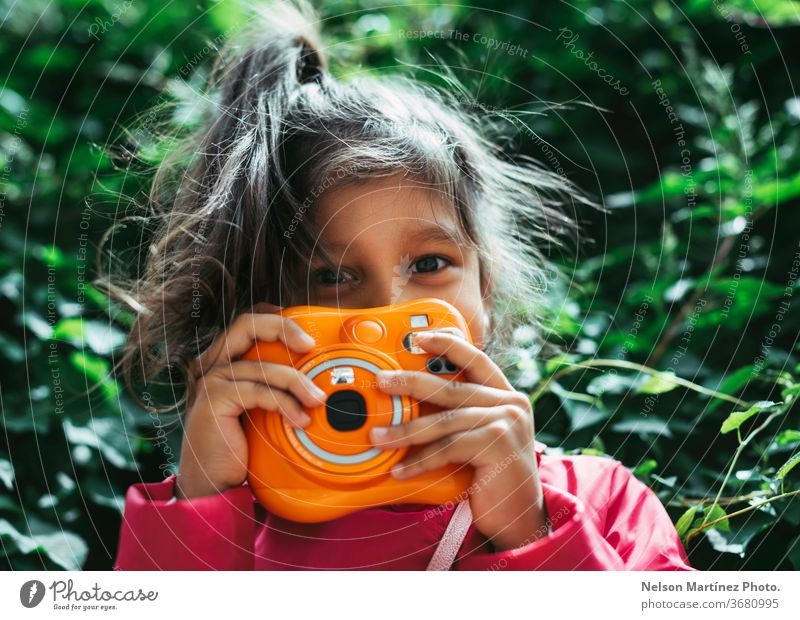 Kleines hispanisches Mädchen mit einer orangefarbenen Instax-Kamera in der Hand. Klassische Kamera. Sie steht vor einem grünen Hintergrund. Kind kleines Mädchen