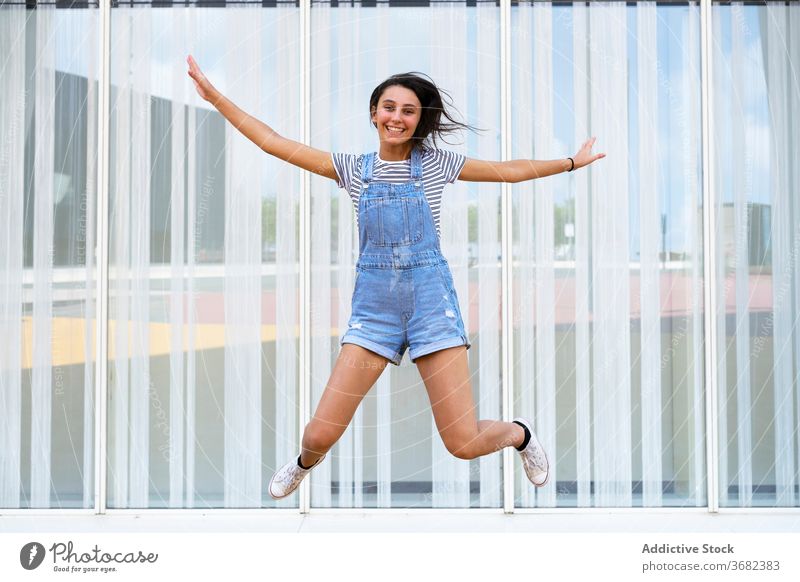 Glückliche Teenager-Mädchen springen in der Nähe von Glaswand Lächeln lässig Sprung Spaß aufgeregt Gebäude Freude froh Optimist modern positiv Moment