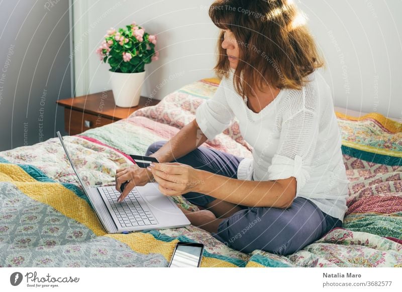Frau, die mit Karte bezahlt, um von zu Hause aus mit dem Laptop auf dem Bett sitzend online einzukaufen. heimwärts Postkarte bezahlen Kauf mittleres Alter
