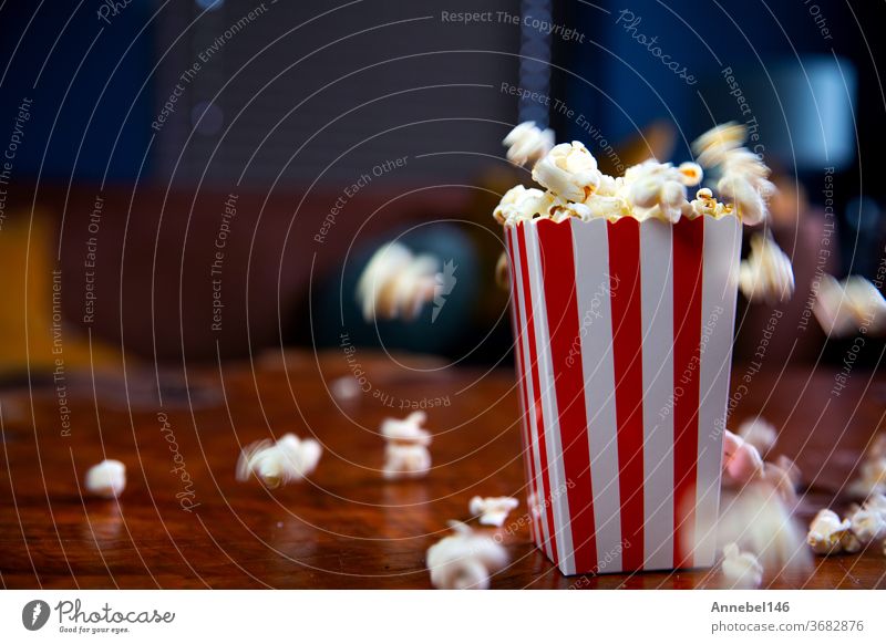 Popcorn fliegt aus Pappkarton. rot-weiß gestreifter Popcorn-Eimer mit fliegendem Popcorn im Wohnzimmer, Film- oder Kinokonzept Popkorn Mais Kasten Snack fallend