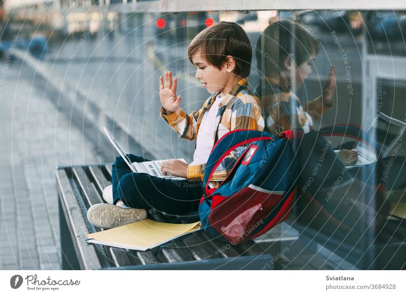 Ein süßer Junge sitzt auf einer Bank, hält einen Laptop auf seinem Schoß, neben einem Rucksack. Der Junge kommuniziert mit Freunden über den Computer und winkt ihnen zu. Bildung, Technologie, Fernunterricht
