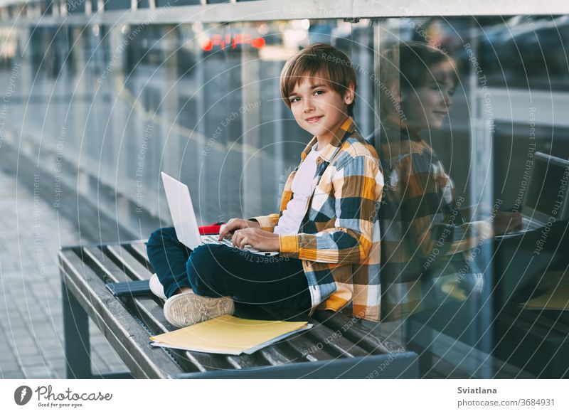 Ein süßer Junge in einem karierten Hemd sitzt auf einer Bank mit einem Laptop und tippt auf der Tastatur, neben einem Rucksack. Der Schüler bereitet sich auf den Unterricht im Freien vor.