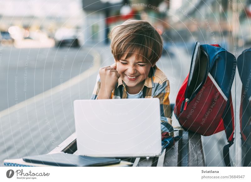 Ein fröhlicher Junge liegt auf einer Holzbank und arbeitet an einem Laptop, neben einem Rucksack. Ein Schüler bereitet sich mit Hilfe des Internets auf den Schulunterricht vor. Soziale Distanz. Fernunterricht