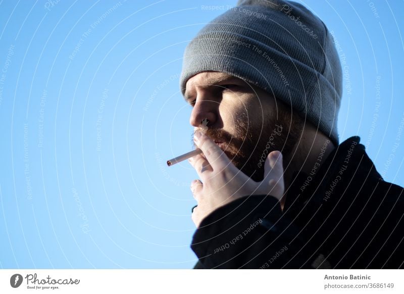Kaukasischer bärtiger Mann mit einer Mütze, der eine Zigarette in der Hand hält, raues Sonnenlicht mit starken Schatten.  Enge Aufnahme mit blauem Himmelshintergrund