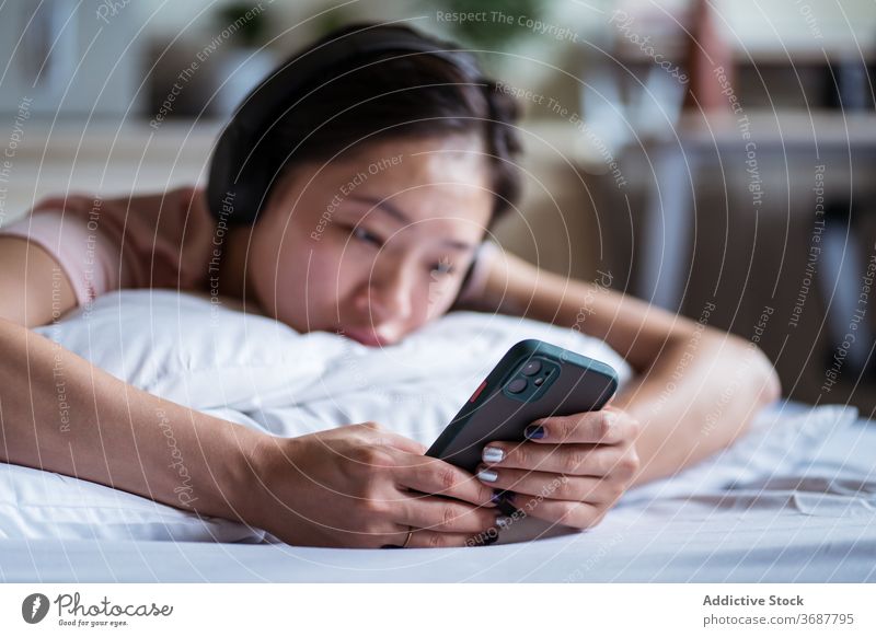 Traurige asiatische Frau, die auf dem Bett auf ihrem Smartphone surft benutzend traurig soziale Netzwerke gemütlich Lügen lesen Schlafzimmer heimwärts