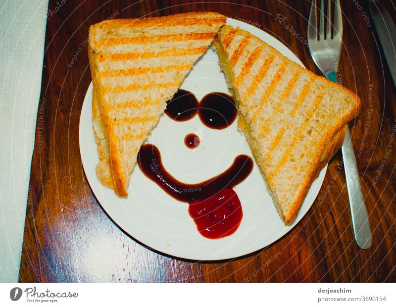 Frühstück Smiley, Ein geteiltes Brot auf dem Teller. Mit Ketschup ein Smiley. Rechts eine Gabel auf einem Holztisch. frühstück snack Lebensmittel Farbfoto