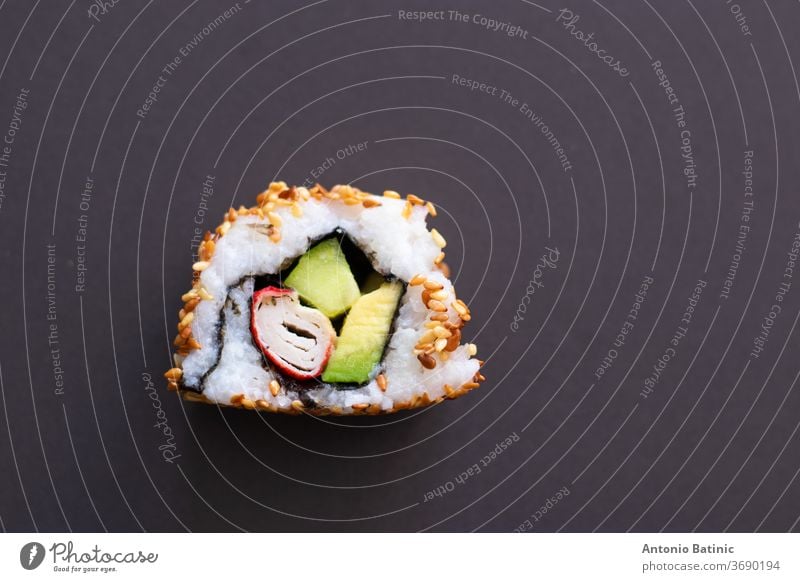 Nahaufnahme eines einzelnen Stückes einer hausgemachten, nicht perfekten Sushi-Rolle. Isoliert auf dunkelschwarzen Hintergrund. Rolle mit Avocado, Surimi, Gurke und gebratenen Sesamkörnern