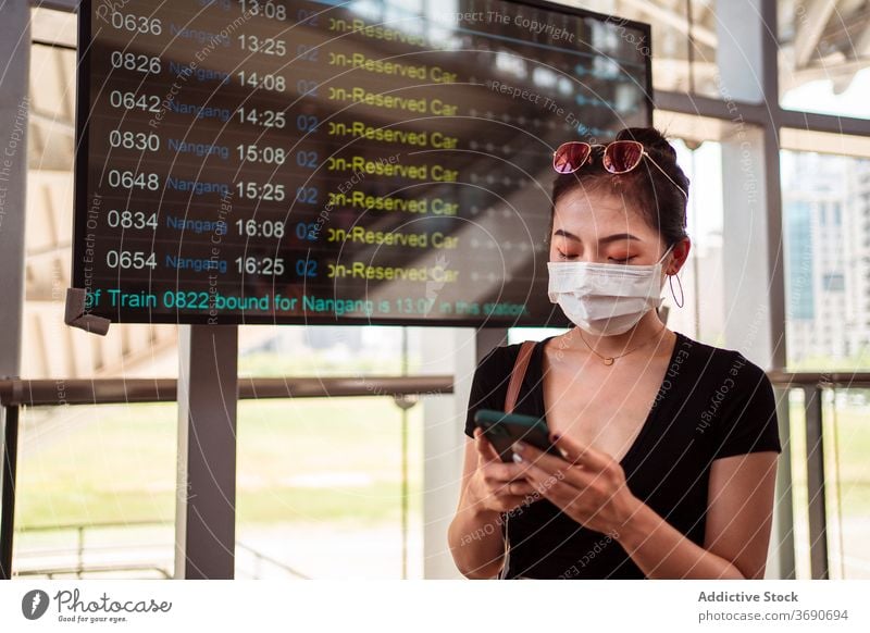 Asiatische Frau benutzt Smartphone am Busbahnhof Station Zeitplan Zeitplanung Mundschutz Coronavirus benutzend warten ethnisch asiatisch digital COVID Fokus
