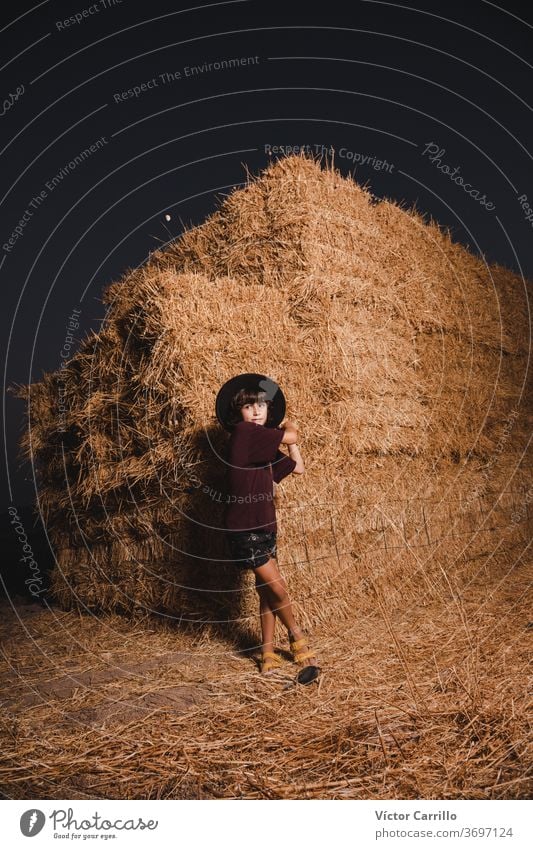 Ein Junge mit Hut, der im Sommer auf den Feldern auf dem Land in einem Strohballenhintergrund spielt Bauernbursche hockend Pflanze Spielen Herbst Kind niedlich