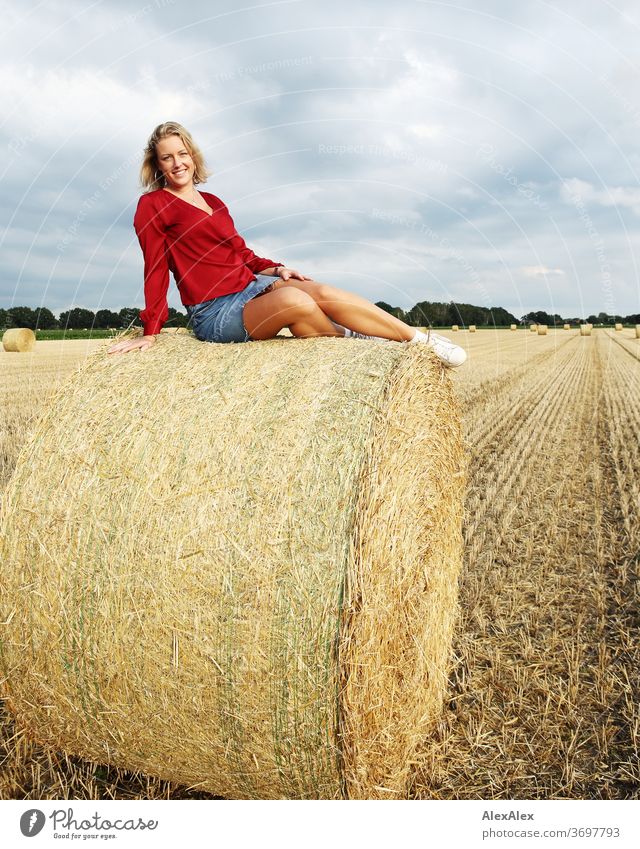 Portrait einer jungen Frau auf einem Strohballen im Abendlicht junge Frau blond Lächeln rot Schmuck schön langhaarig Landschaft Wolken Himmel gebräunt