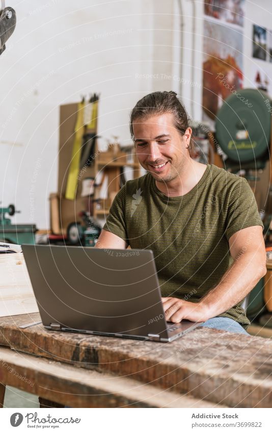 Lächelnde männliche Holzarbeiter mit Laptop in der Werkstatt Holzarbeiten Zimmerer Mann Projekt Arbeit benutzend Hobelbank Inhalt Netbook modern Gerät