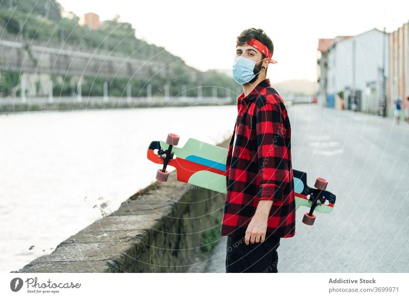 Männlicher Hipster in medizinischer Maske in der Stadt Skater Longboard Spazierweg Mundschutz Mann Coronavirus Spaziergang behüten männlich chirurgisch jung
