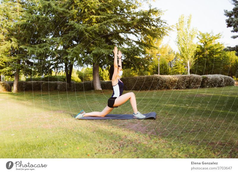 Junge brünette Frau mit Maske, die sich auf einer Matte im Park bewegt und streckt Mundschutz passen Yoga Pandemie COVID Fitness Übung Meditation Coronavirus